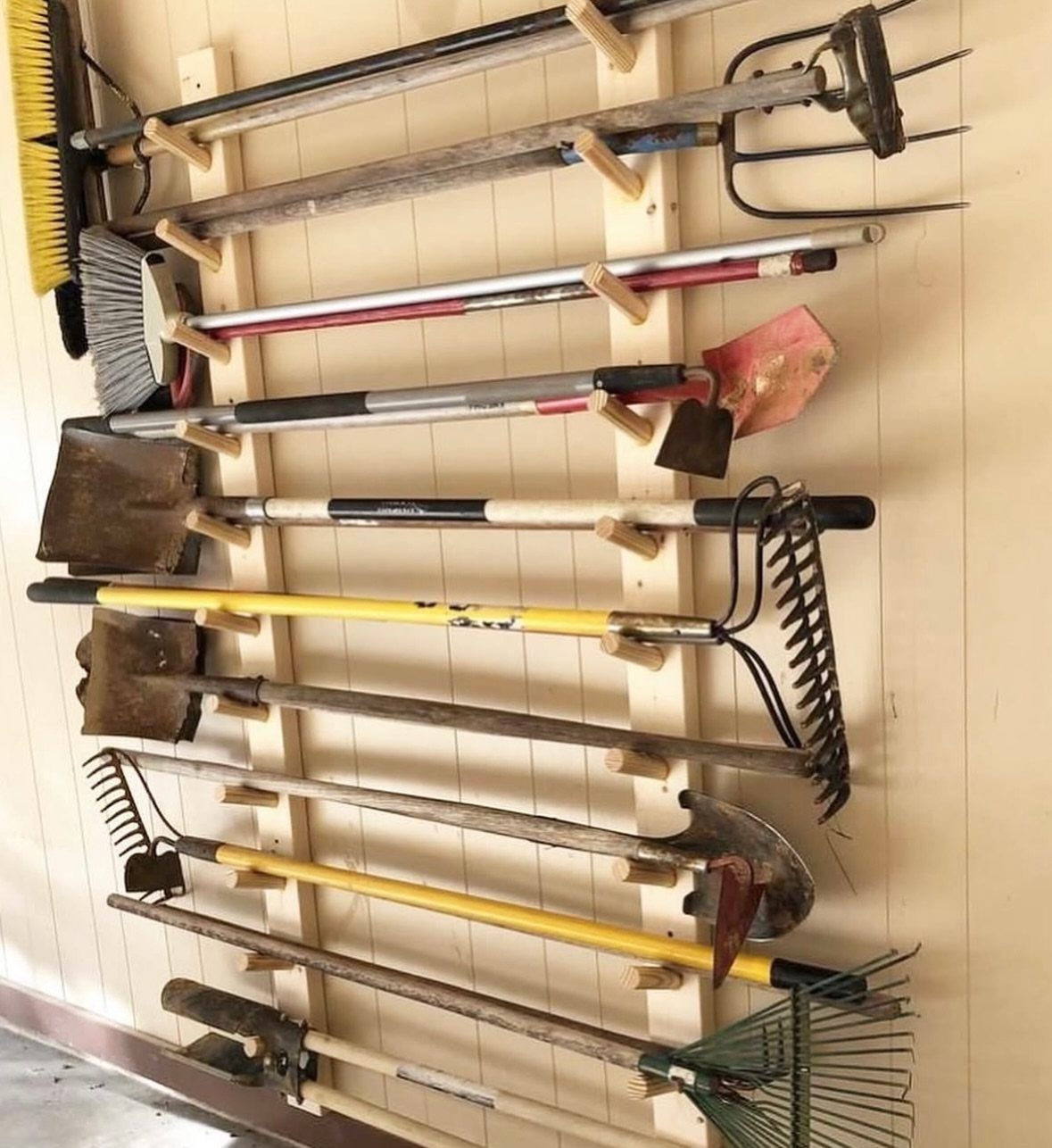 Long-handled garden tools hanging organized on a wooden wall rack in a garage.