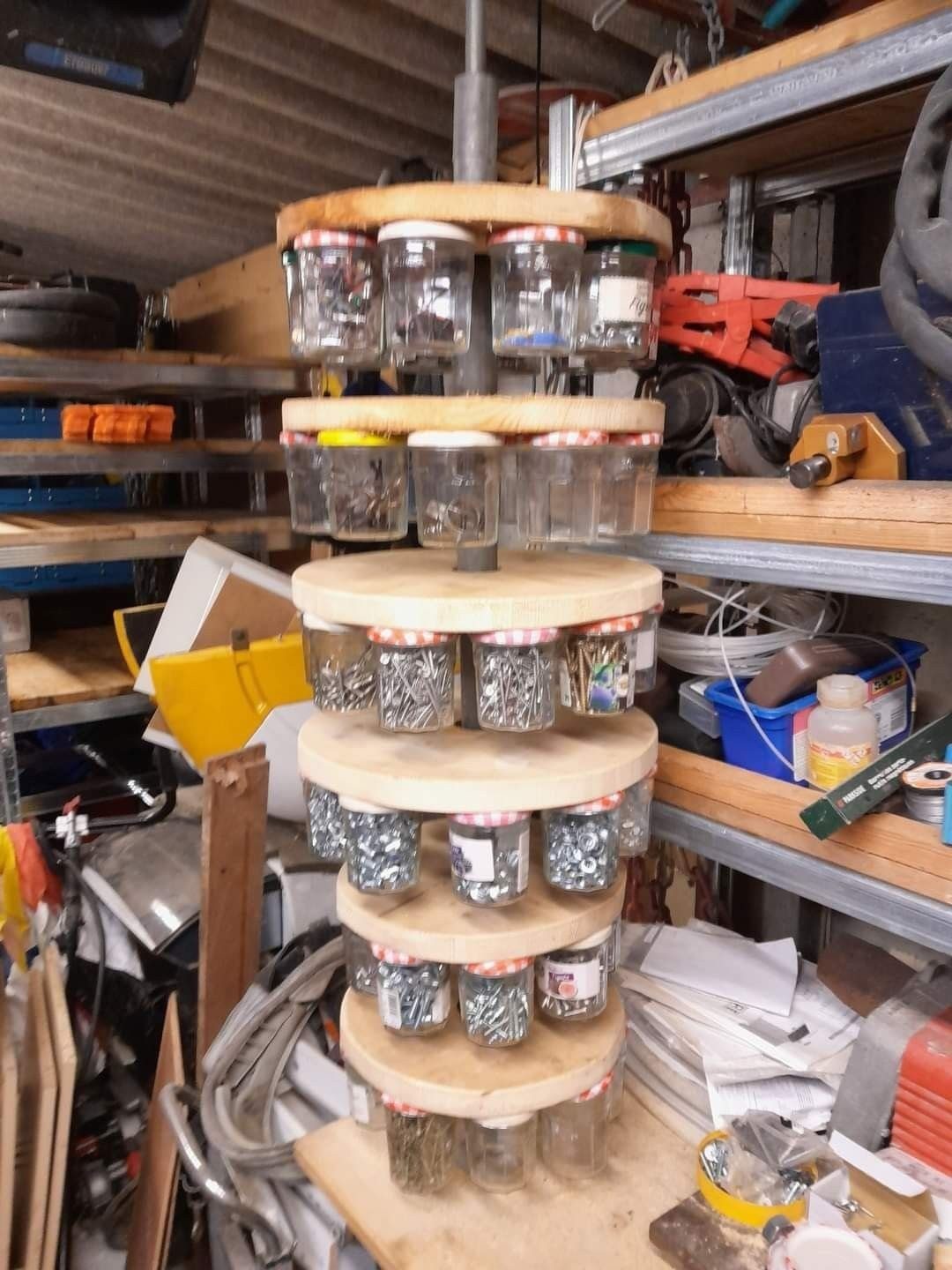 A wooden tiered organizer holding glass jars filled with various metal screws and nails in a workshop.